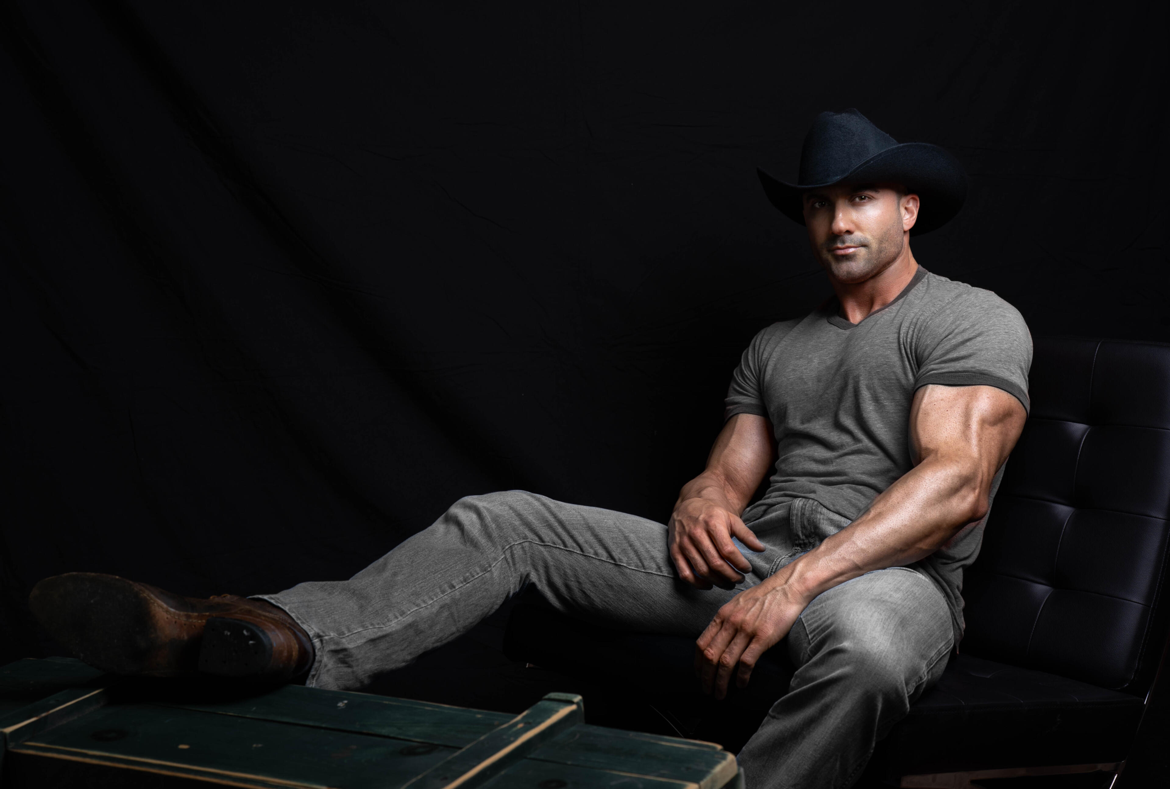 Professional modelling photography of Tom Rooney - performer, author, entrepreneur - great picture, sitting in black leather chair with cowboy boots, jeans, T-shirt, and a cowboy hat